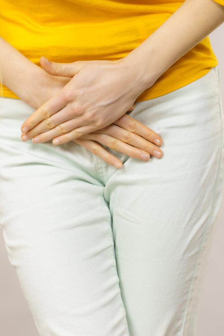 Eine Person hält sich mit beiden Händen den Unterbauch und trägt ein gelbes Oberteil sowie eine helle Hose.