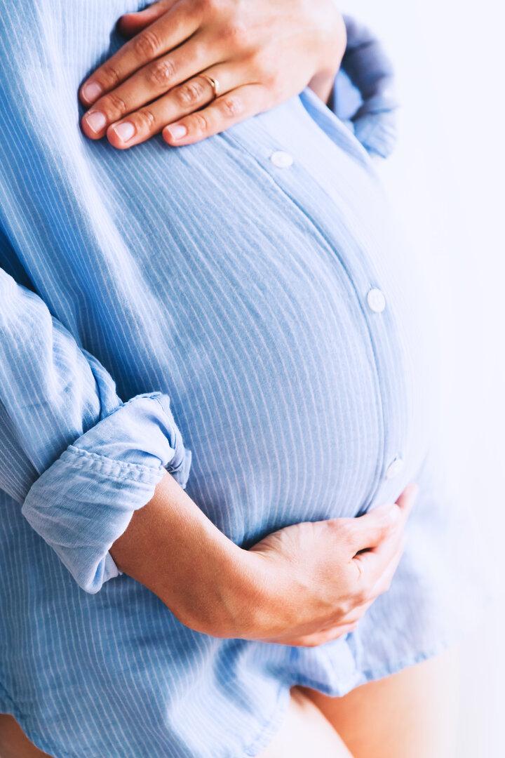 Eine schwangere Frau in einem hellblauen Hemd hält ihren Bauch.