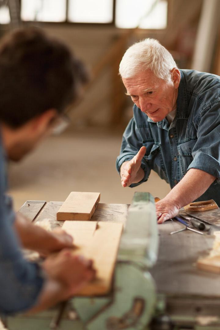 Ein älterer Mann erklärt einem jüngeren Mann in einer Werkstatt die Holzbearbeitung an einer Werkbank.