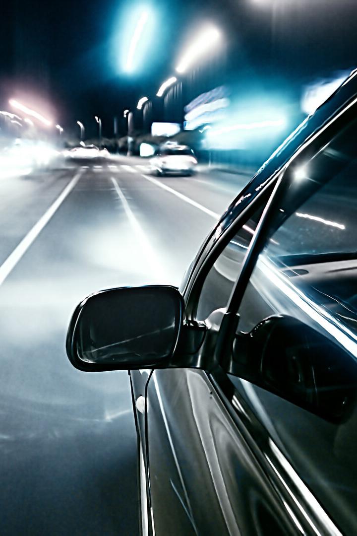 Ein Auto fährt nachts mit hoher Geschwindigkeit auf einer beleuchteten Straße, Lichter ziehen Spuren.