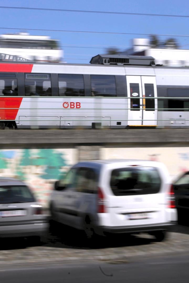 Ein roter und silberner ÖBB-Zug fährt an parkenden Autos vorbei.