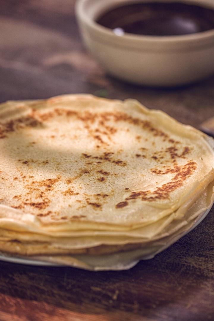 Ein Stapel Palatschinken liegt auf einem Teller, daneben stehen Schalen mit Saucen auf einem Holztisch.