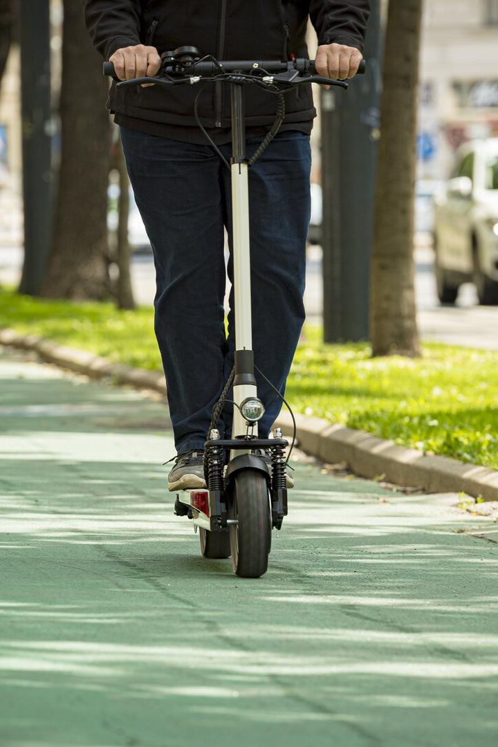 Eine Person fährt mit einem E-Scooter und eine weitere mit dem Fahrrad auf einem grünen Radweg entlang einer Baumallee.