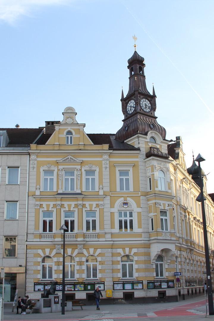 Historisches gelbes Gebäude mit Uhrturm und mehreren Flaggen davor an einem Platz bei Tageslicht.