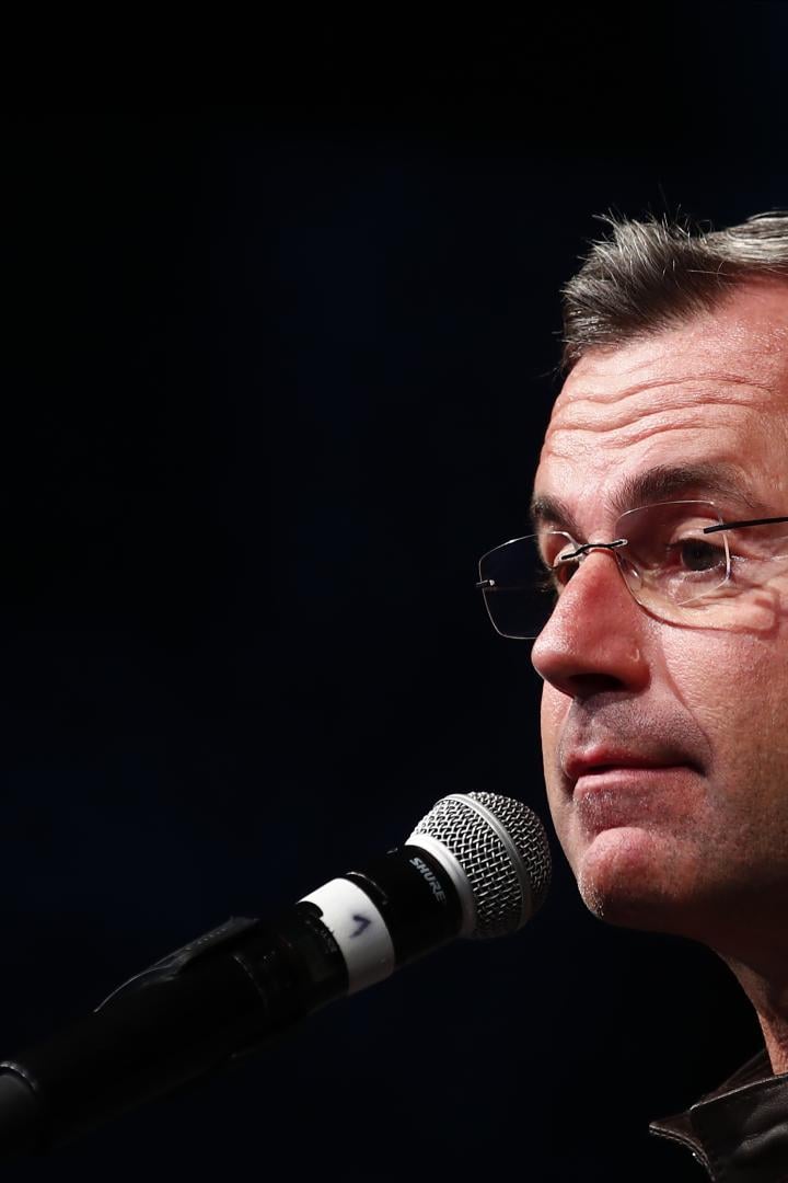 Ein Mann mit Brille und grauen Haaren spricht in ein Mikrofon, dunkler Hintergrund.