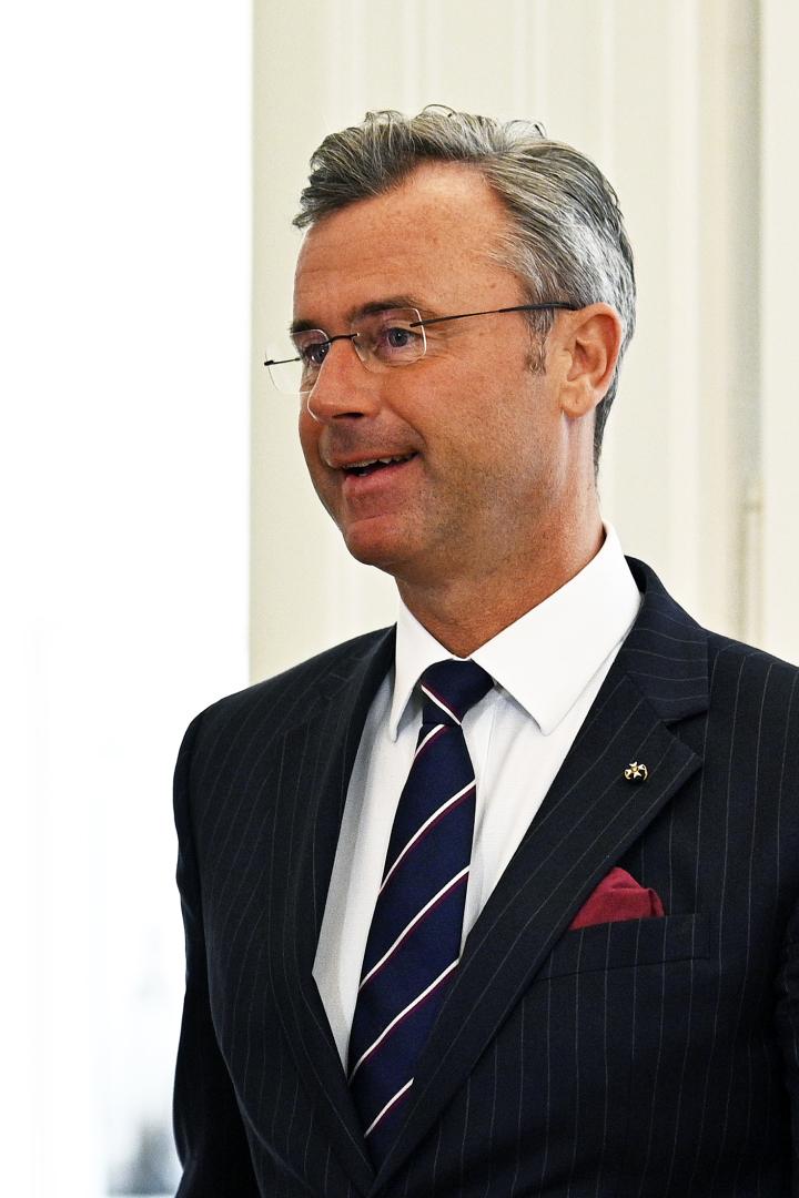 Ein Mann mit Brille trägt einen dunkel gestreiften Anzug, ein weißes Hemd und eine gestreifte Krawatte.