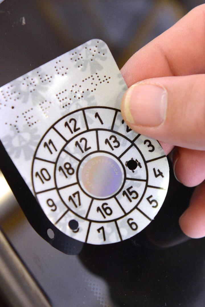 Eine Hand klebt eine österreichische Prüfplakette an eine Windschutzscheibe.