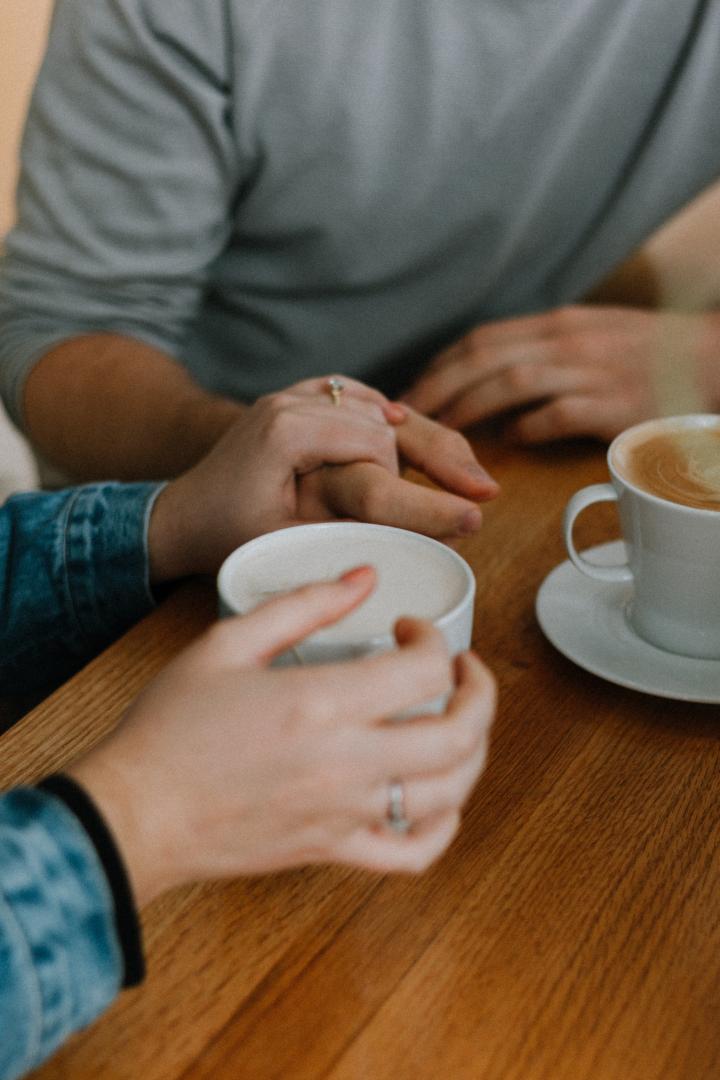 Zwei Personen sitzen an einem Holztisch, halten Händchen und trinken gemeinsam Kaffee.