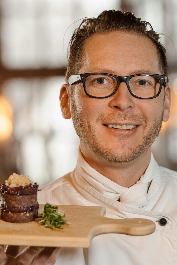 Ein Koch in weißer Jacke hält ein Brett auf dem Fleisch gestapelt und mit Kräutern verziert liegt.