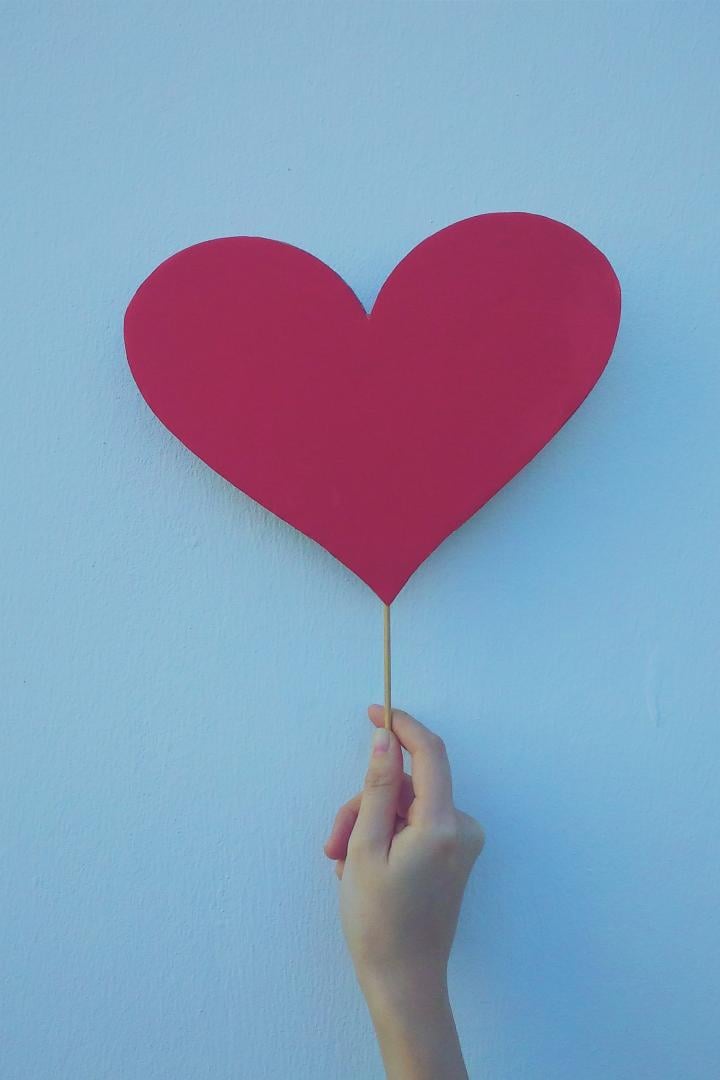 Eine rosa Herz auf einem Holzstiel wird von einer Hand an eine blaue Wand gehalten.