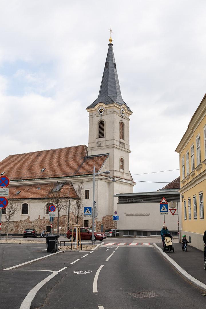 Eine Kirche mit hohem Turm steht an einer Straße, umgeben von alten Gebäuden und mehreren Fußgängern.