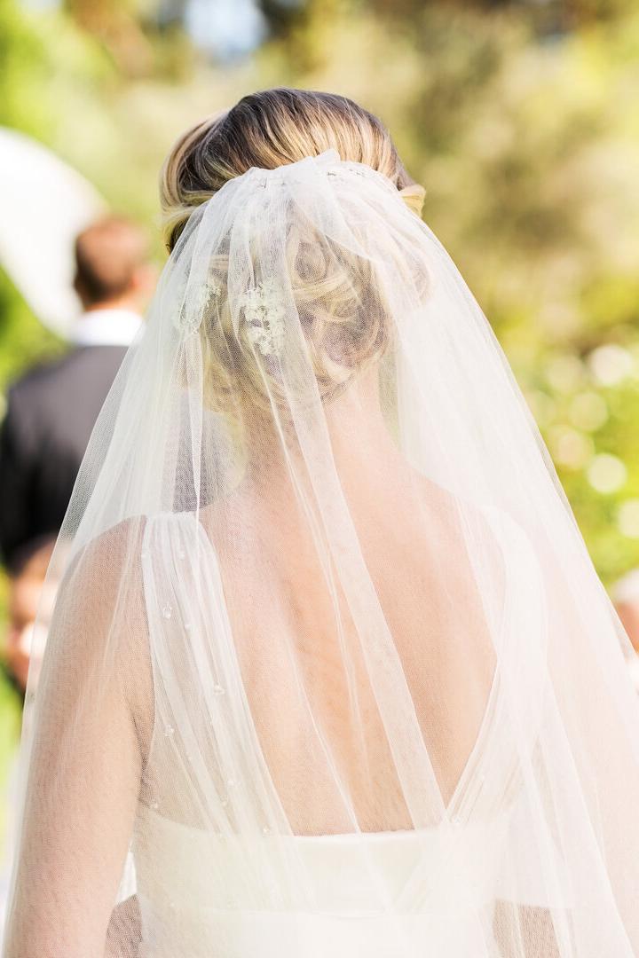 Eine Braut mit Schleier steht im Freien vor einem Traubogen, während Gäste und ein Redner warten.