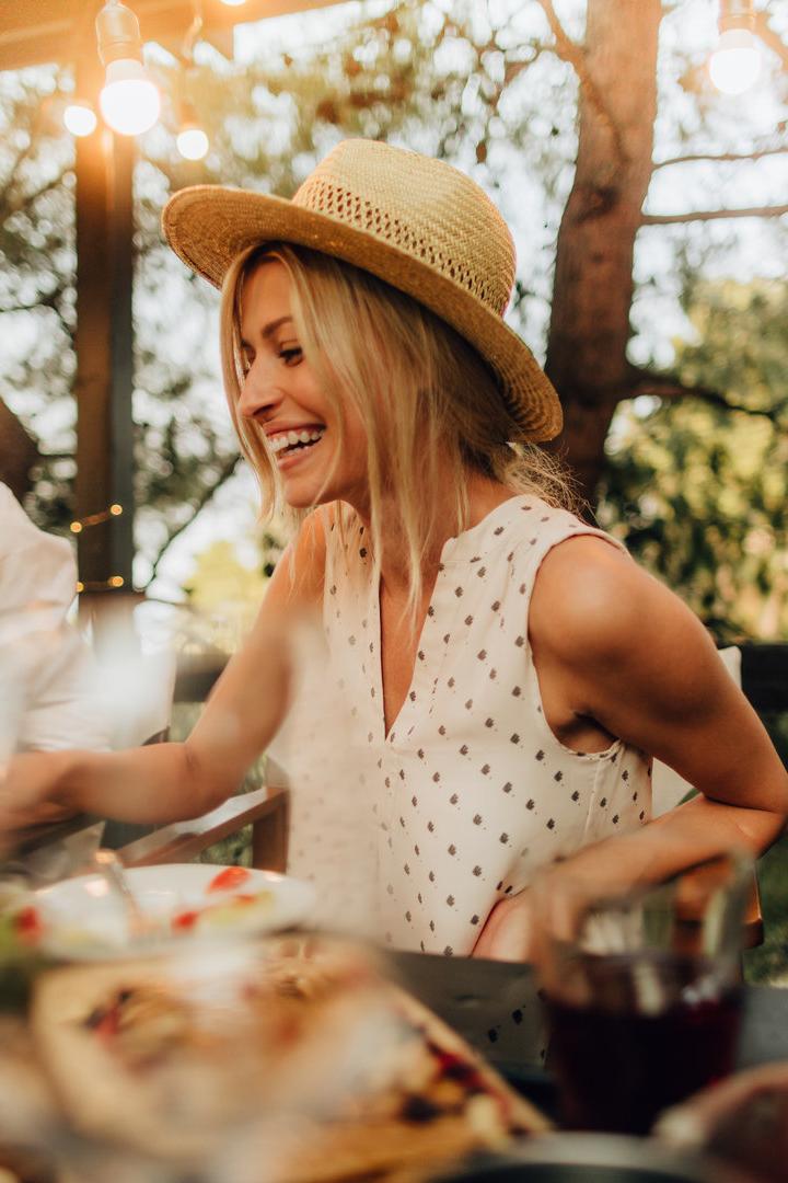 Eine lachende Frau mit Hut sitzt mit einem Mann bei einem Abendessen im Freien.