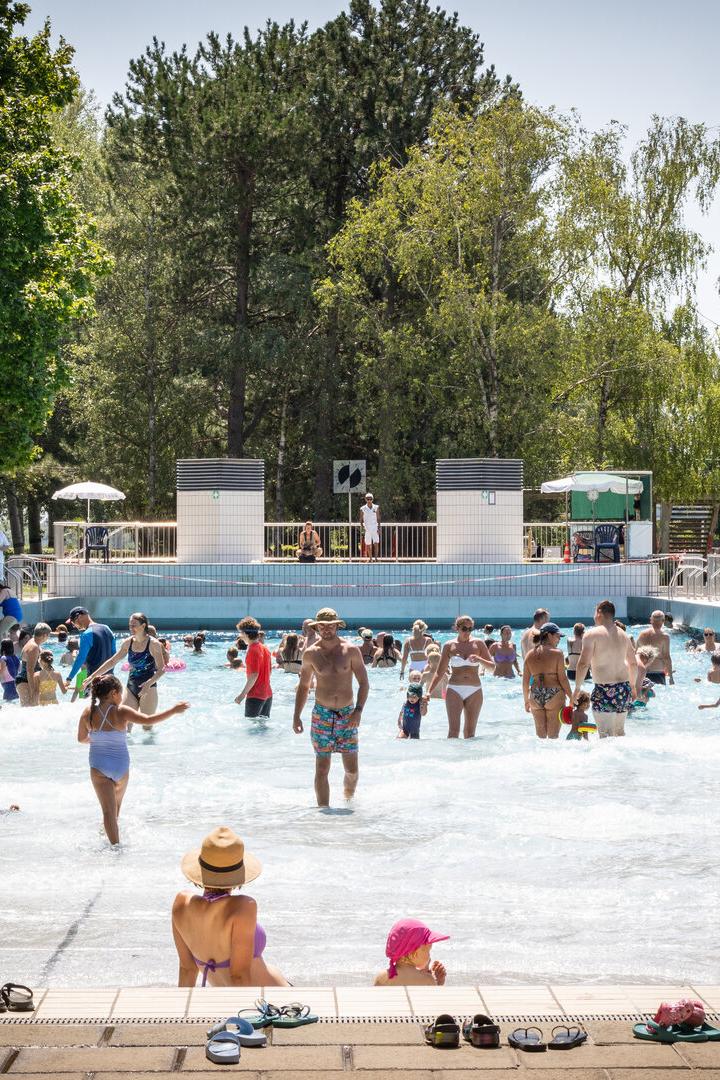 Viele Menschen, darunter Kinder und Erwachsene, genießen einen sonnigen Tag in einem Freibad mit flachem Wasser und umgebenden Bäumen.