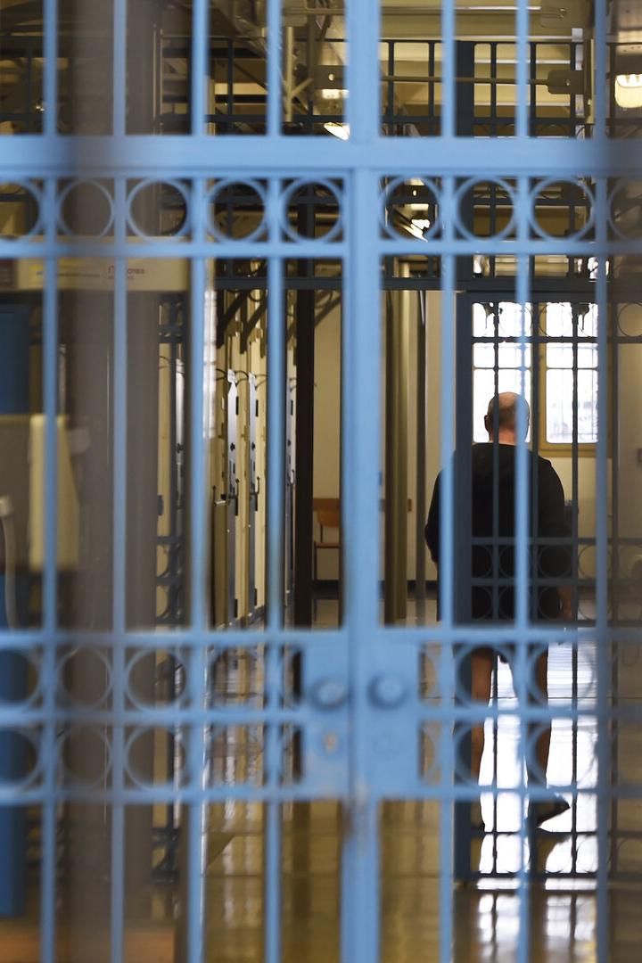 Eine Person steht hinter blauen Gitterstäben in einem hellen Flur mit mehreren Türen und Fenstern.