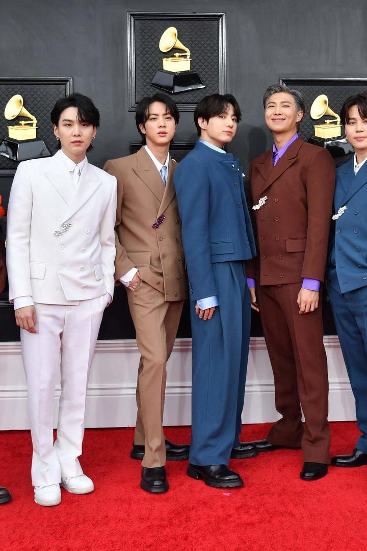 Sieben südkoreanische Männer in roten, weißen, beigen und blauen Anzügen auf einem roten Teppich. Im Hintergrund sieht man eine schwarze Wand mit mehreren goldenen Preisen in Form eines alten Plattenspielers.