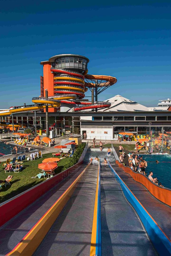 Blick von einer Wasserrutsche auf ein belebtes Schwimmbad mit Rutschen und Liegewiese.