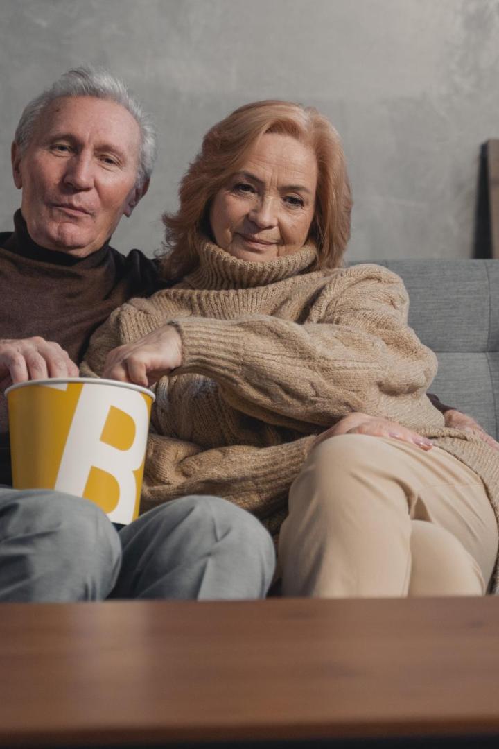 Ein älteres Paar sitzt mit einer Popcorntüte auf einem Sofa.