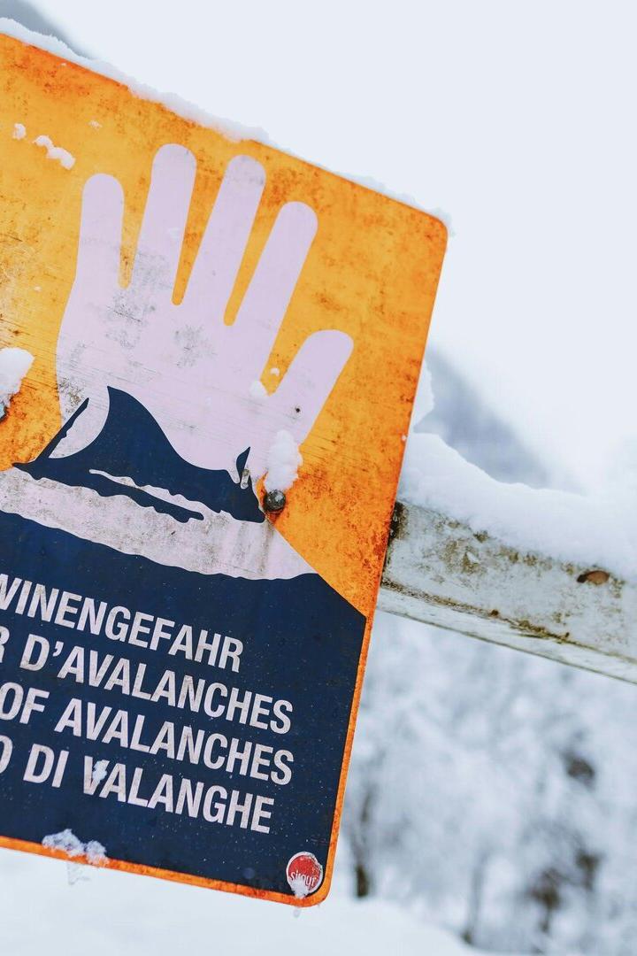 Warnschild mit erhobener Hand und Hinweis auf Lawinengefahr in mehreren Sprachen, umgeben von verschneiter Landschaft.