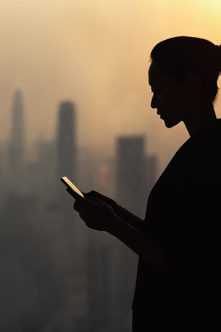 Silhouette of young woman using smartphone next to window with cityscape