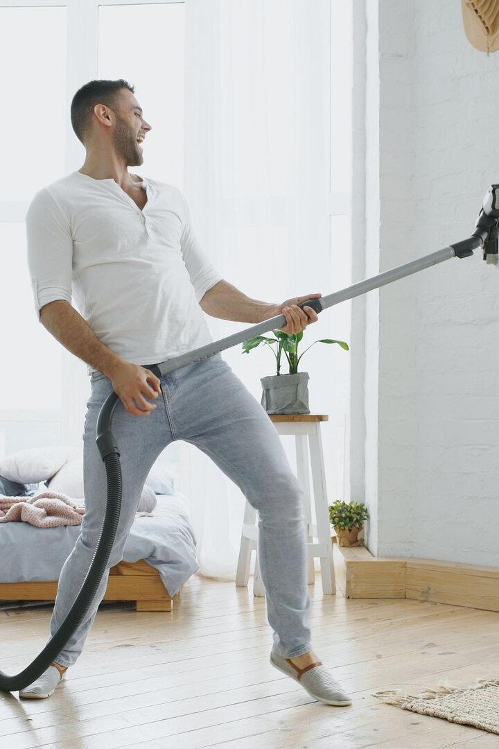 Young man having fun cleaning house with vacuum cleaner dancing like guitarist