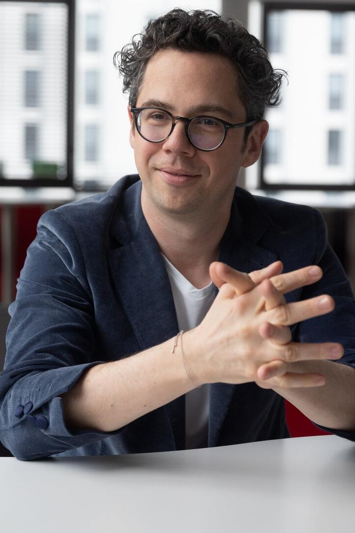 Ein Mann mit Brille und lockigen Haaren sitzt lächelnd an einem Tisch in einem modernen Büro.