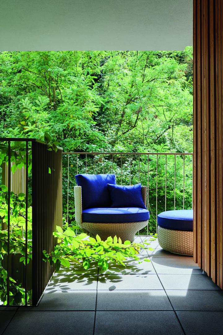 Ein Sessel und ein Hocker mit blauen Kissen stehen auf einem Balkon mit Blick auf üppiges Grün.