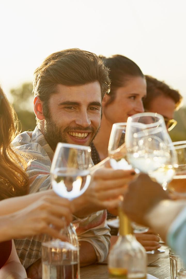 Freunde stoßen lachend bei einem sonnigen Essen im Freien mit Weingläsern an.