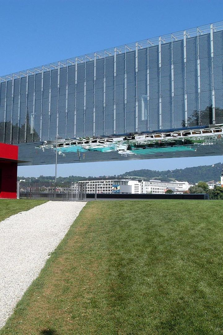 Ein moderner, verglaster Bau mit spiegelnder Fassade und einem kleinen roten Kubus steht auf einer grünen Wiese.