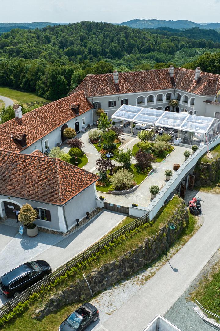 Luftaufnahme eines großen Gutshofs mit roten Dächern, Innenhof und Terrassen-Zelt, umgeben von grünen Hügeln.