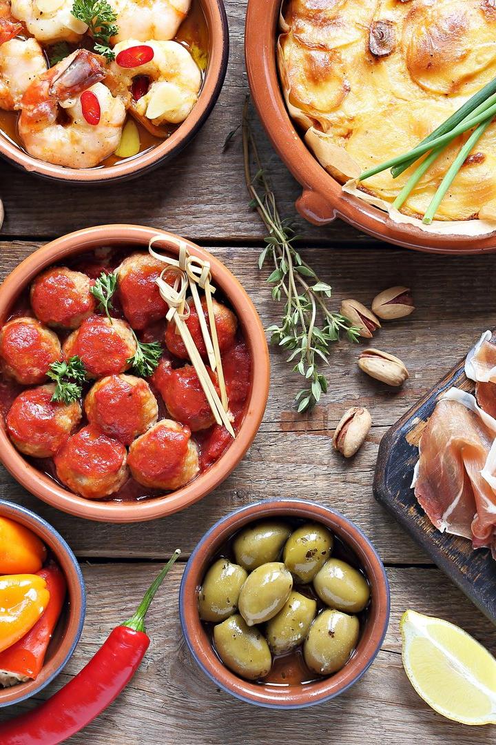 Verschiedene Tapas, darunter Oliven, Fleischbällchen, Käse und Kartoffelgratin, sind auf einem Holztisch angerichtet.