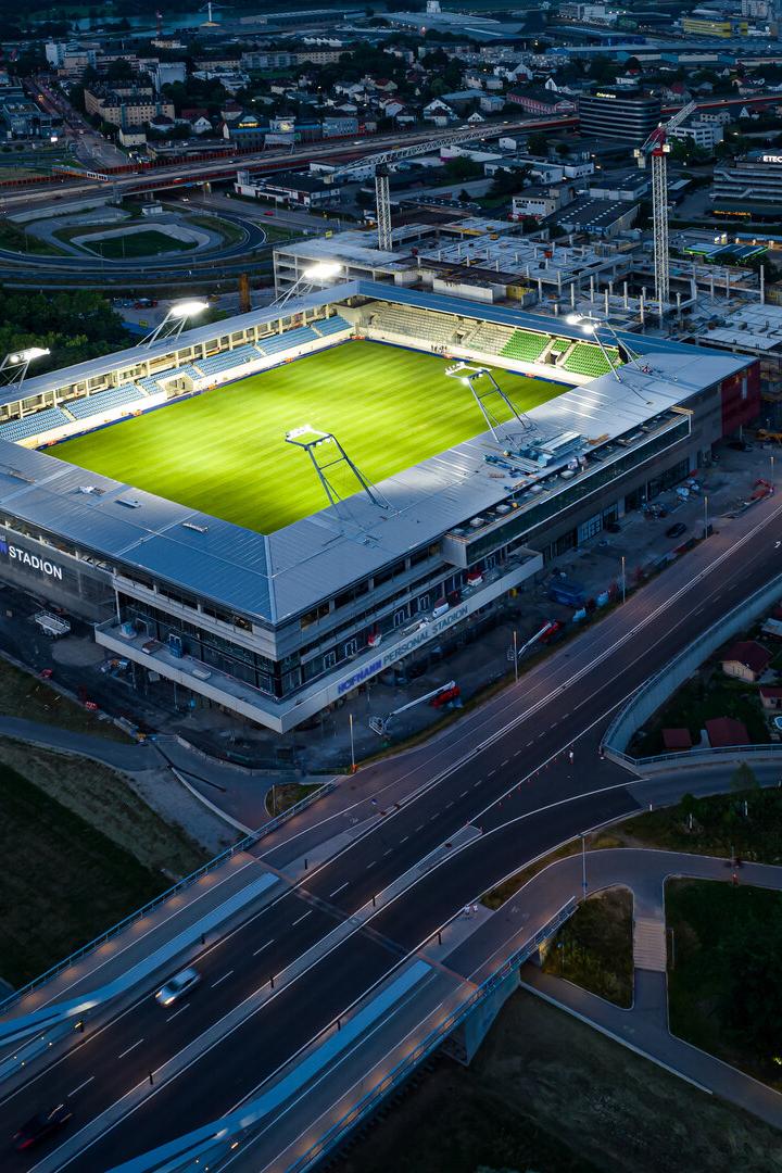 Ein beleuchtetes Fußballstadion mit grünem Spielfeld liegt an einer großen Straßenkreuzung in einer Stadt bei Nacht.
