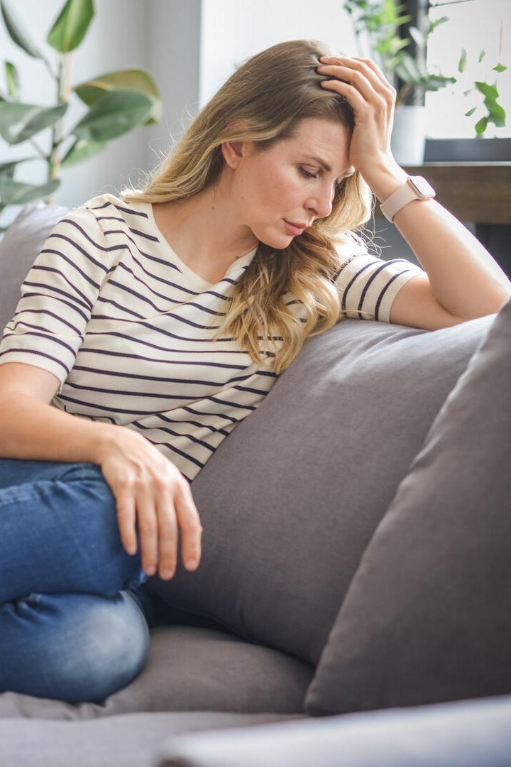 Eine Frau mit blonden Haaren sitzt nachdenklich auf einem grauen Sofa.