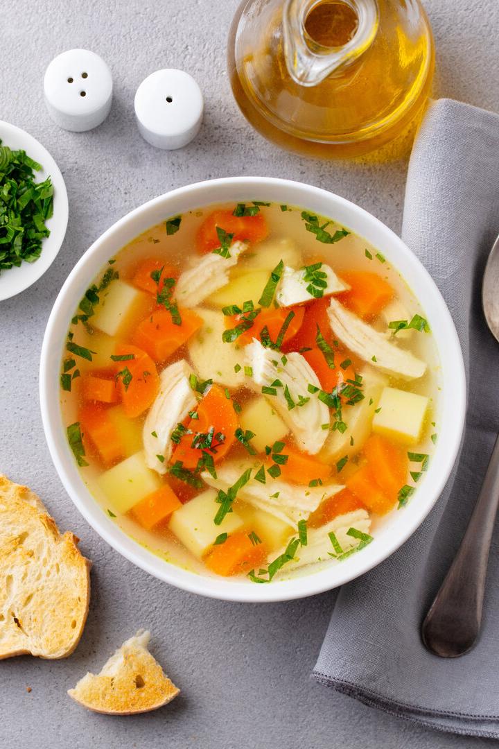 Eine Schale Hühnersuppe mit Gemüse und frischen Kräutern, daneben Brot, Kräuter und ein Löffel auf Serviette.