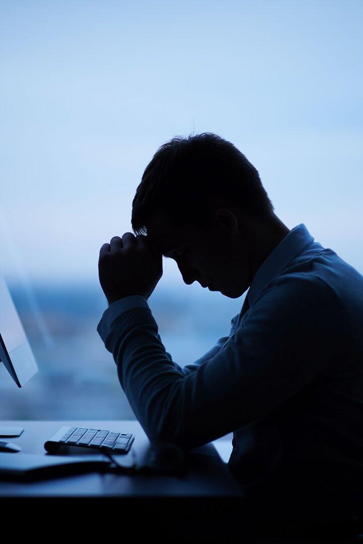 Eine Person sitzt mit gesenktem Kopf und verschränkten Händen nachdenklich vor einem Computer am Schreibtisch.