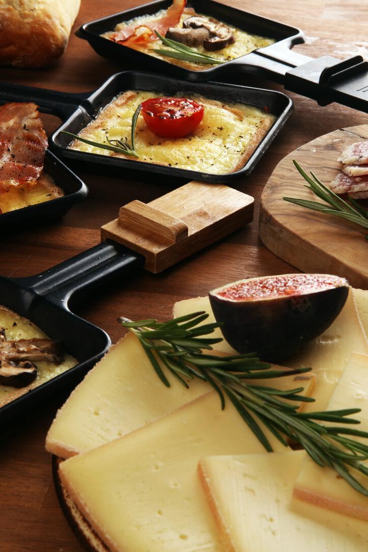 Mehrere Raclettepfännchen mit geschmolzenem Käse, Gemüse und Speck, dazu Käse- und Wurstscheiben mit Rosmarin.