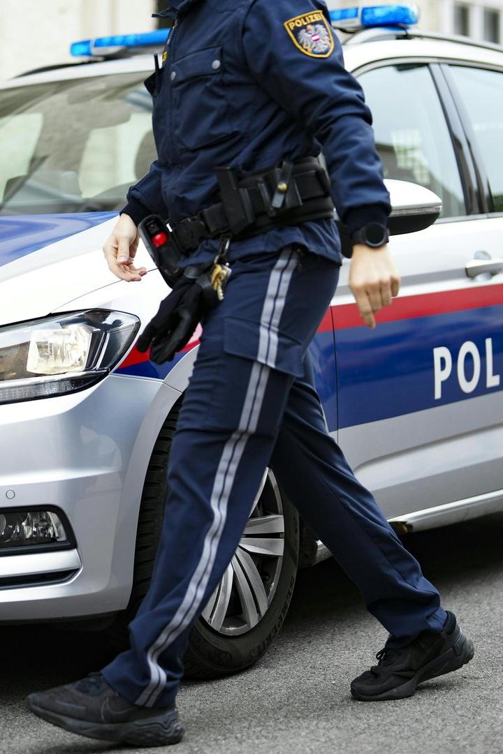 Ein Polizist in Uniform geht an einem silbernen Polizeiauto mit blauen und roten Streifen vorbei.