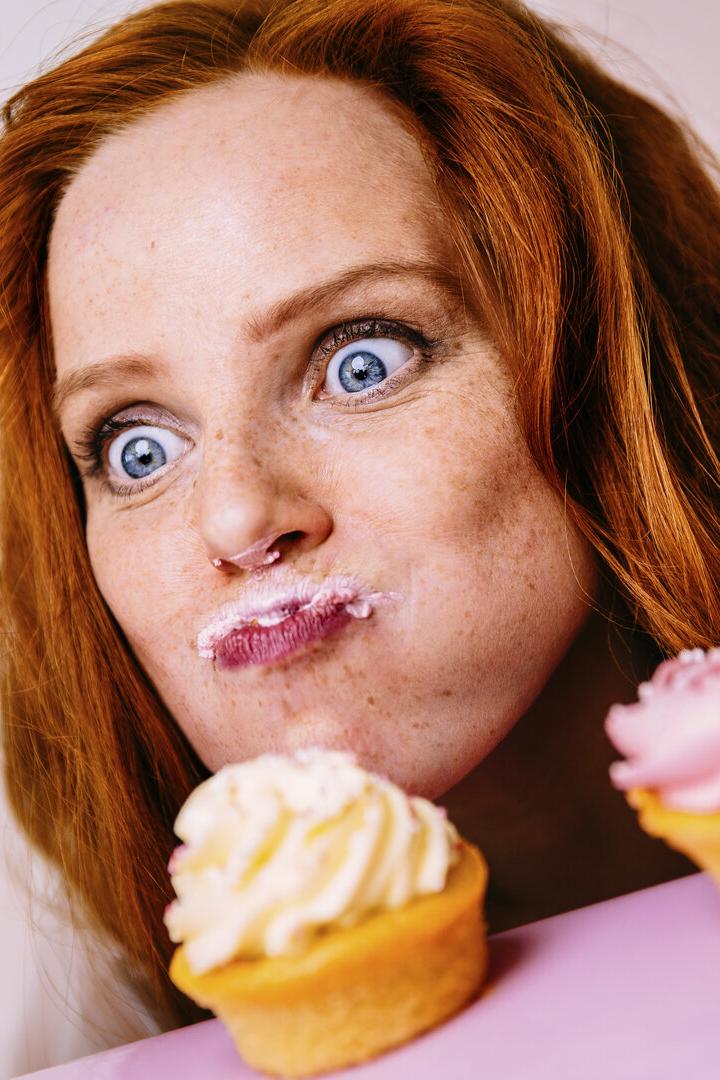 Eine rothaarige Frau mit Zuckerguss im Gesicht hält einen Cupcake und blickt mit großen Augen auf weitere Cupcakes.