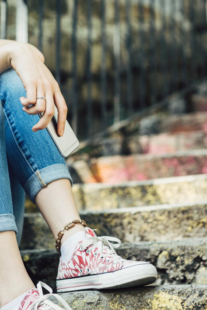 Eine junge Frau sitzt mit Handy auf einer Treppe, gekleidet in Jeans und gelbem Oberteil.
