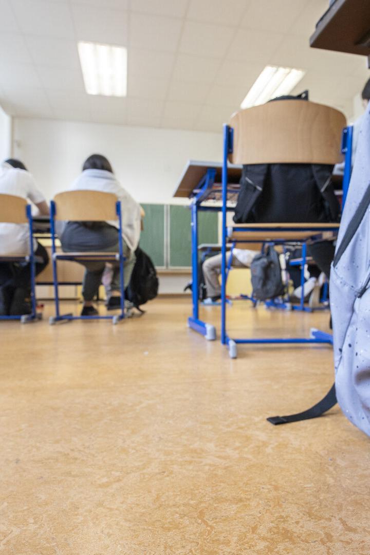 Ein hellgrauer Rucksack steht im Vordergrund eines Klassenzimmers.