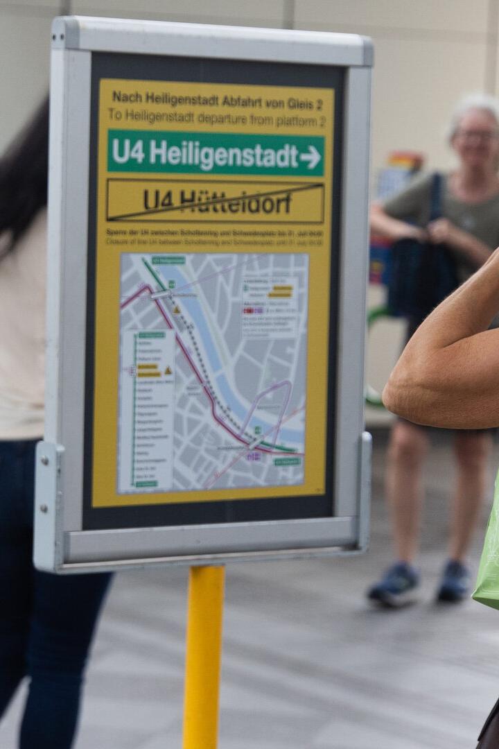 Eine Frau mit grüner Einkaufstasche betrachtet einen U-Bahn-Plan an einer Station, während andere Personen vorbeigehen.