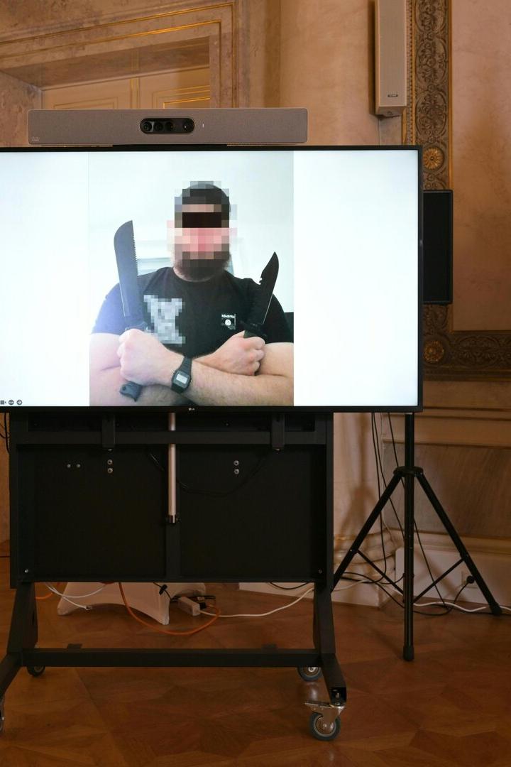 Auf einem Bildschirm ist ein Mann mit zwei Messern zu sehen; im Hintergrund die Europa- und Österreich-Flagge.