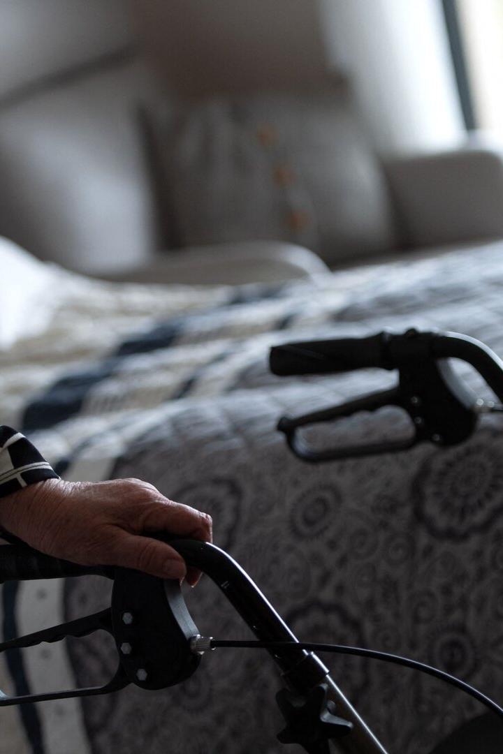 Eine ältere Frau stützt sich auf einen Rollator in einem Schlafzimmer.