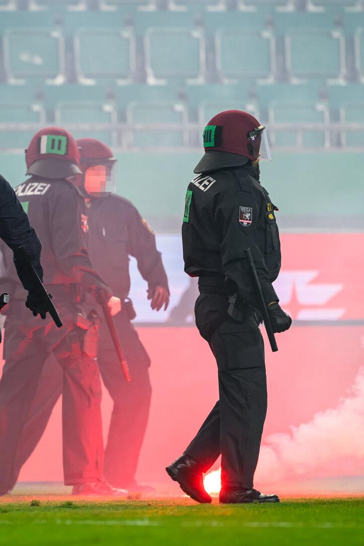 Polizisten in Schutzausrüstung stehen auf einem Fußballfeld inmitten von rotem Rauch.