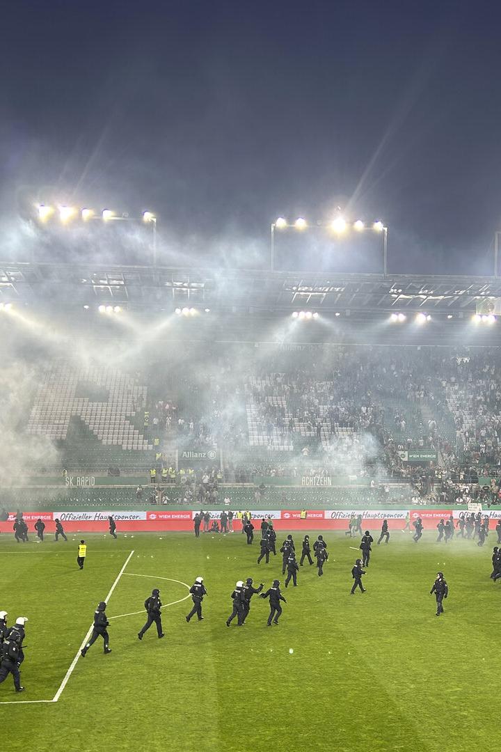 Polizeibeamte betreten ein Fußballfeld, das mit Rauch gefüllt ist, während Fans auf den Tribünen sitzen.
