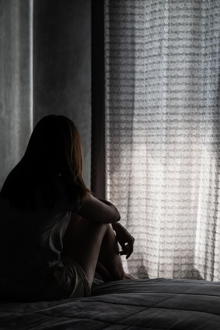 Eine Person sitzt nachdenklich auf einem Bett und blickt auf geschlossene Vorhänge, durch die Licht fällt.