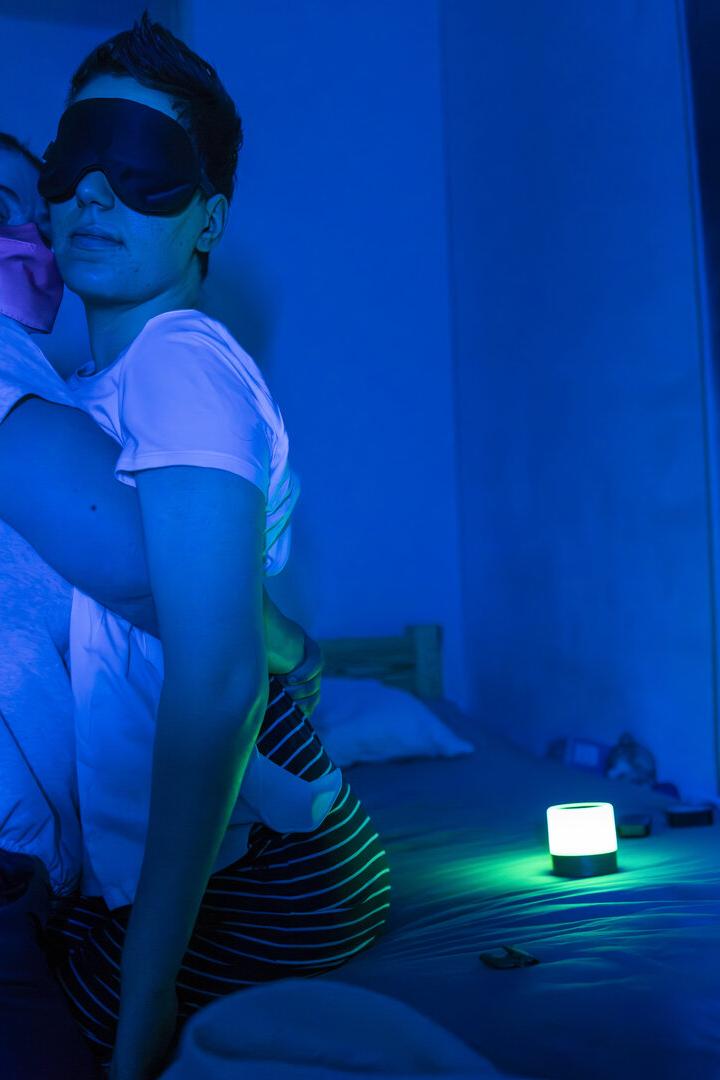 Eine Frau mit Maske umarmt eine Frau mit Schlafmaske in einem blau beleuchteten Raum.