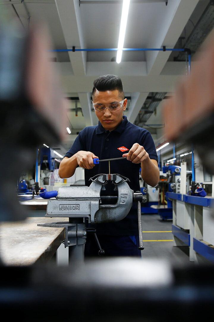 Ein Mann mit Schutzbrille arbeitet mit einer Feile an einem Werkstück, das in einem Schraubstock eingespannt ist.