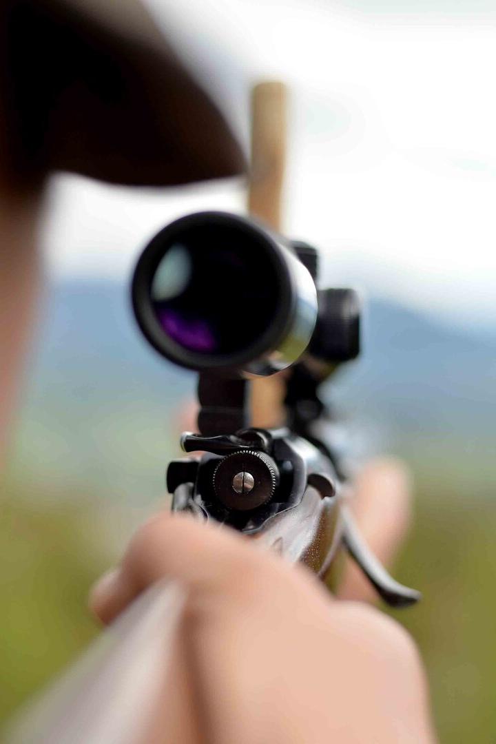 Eine Person zielt mit einem Gewehr samt Zielfernrohr in eine unscharfe Landschaft.