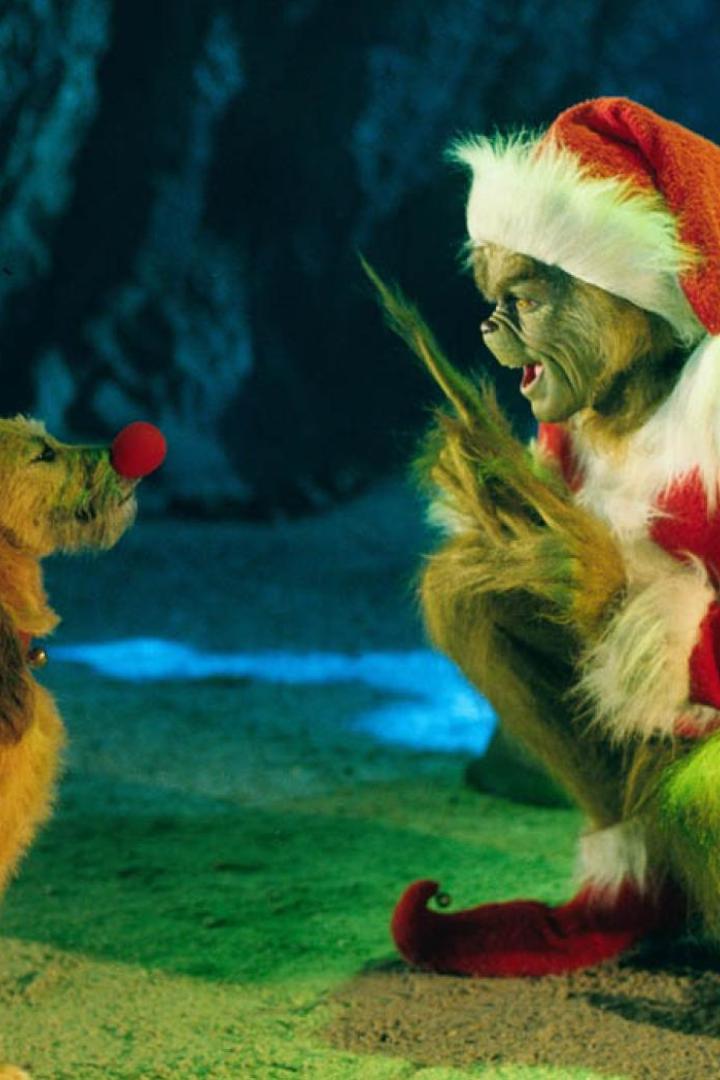 Der Grinch im Weihnachtsmannkostüm hockt vor einem Hund mit roter Nase und Geweih.